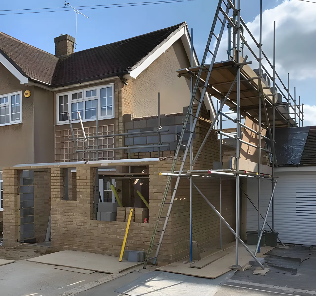 Construction scaffolding set up around a two-story house with brickwork being added to the lower level, under a partly cloudy sky.
