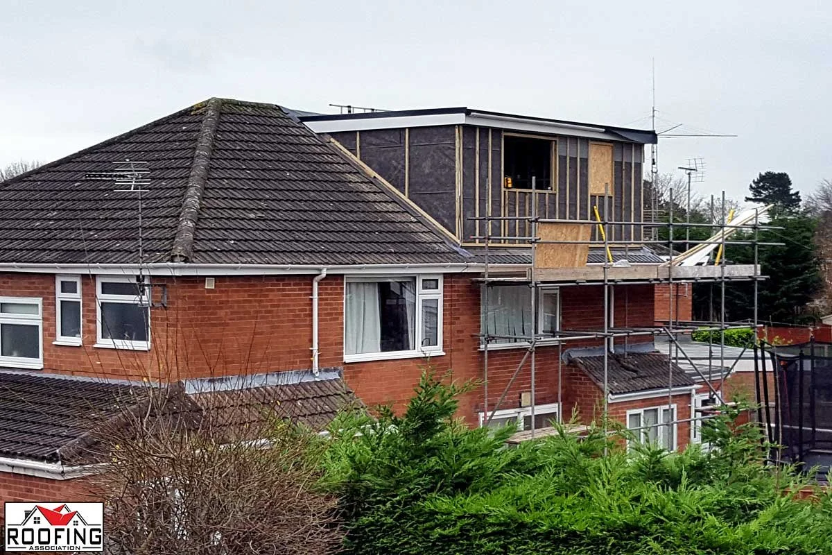 A house with a brick exterior and a tiled roof under construction, featuring scaffolding on the side and a new upper level being added.