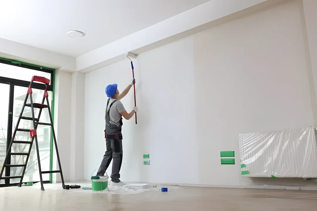 A person painting a wall with a roller brush in a room, wearing a safety helmet and grey overalls, with a ladder nearby and painting supplies on the floor.