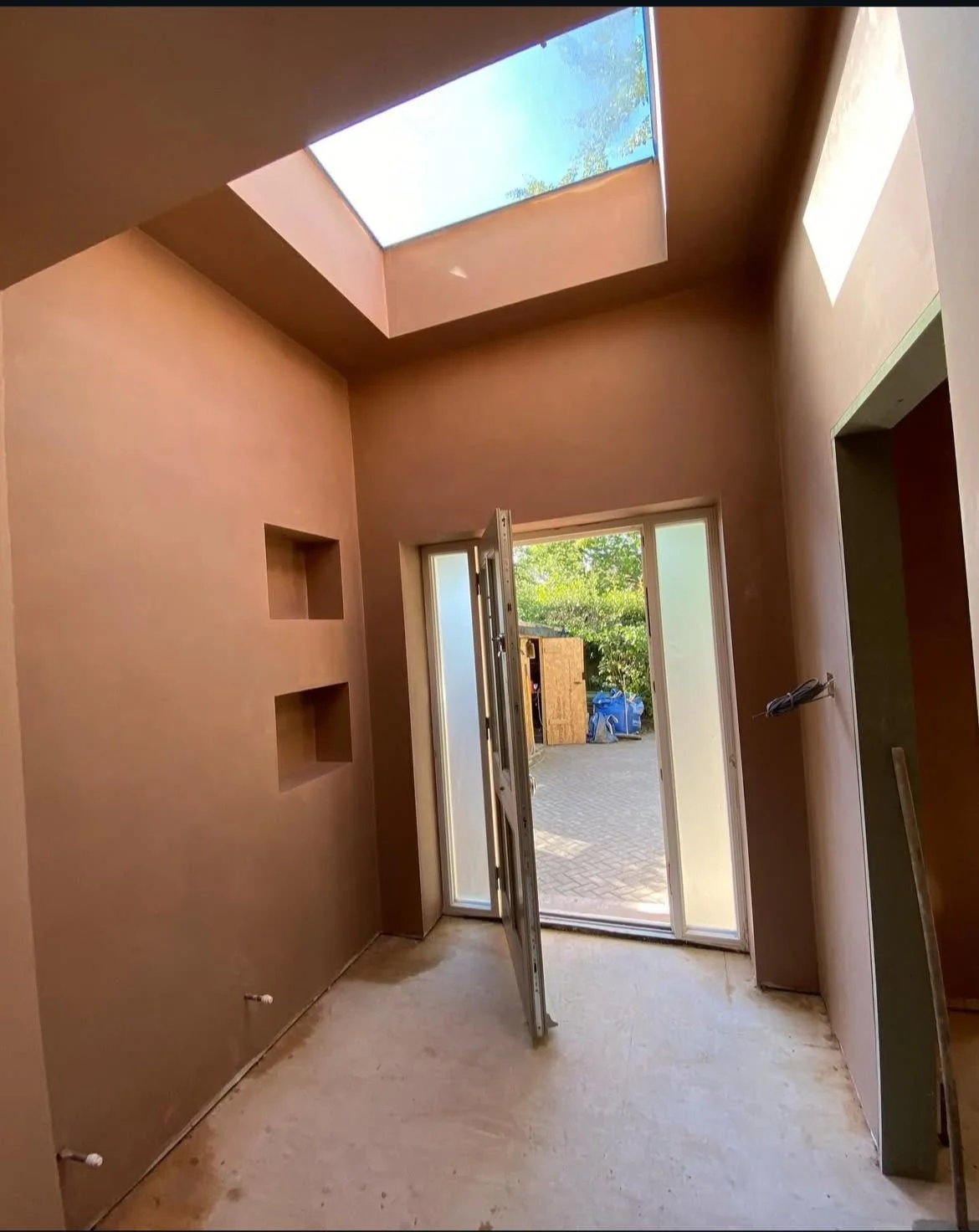 Interior of a room under construction with brown walls and a large skylight ceiling, an open door leading outside, and built-in wall niches.
