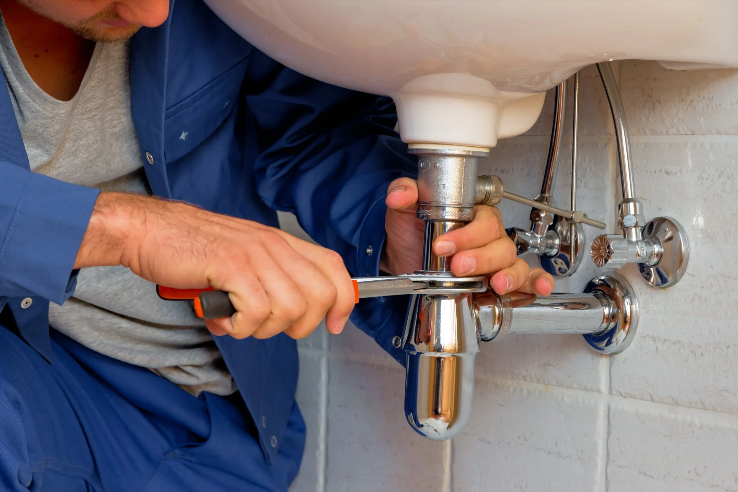 A plumber wearing a blue uniform and gray shirt working underneath a kitchen sink, tightening a pipe or fitting with a wrench.